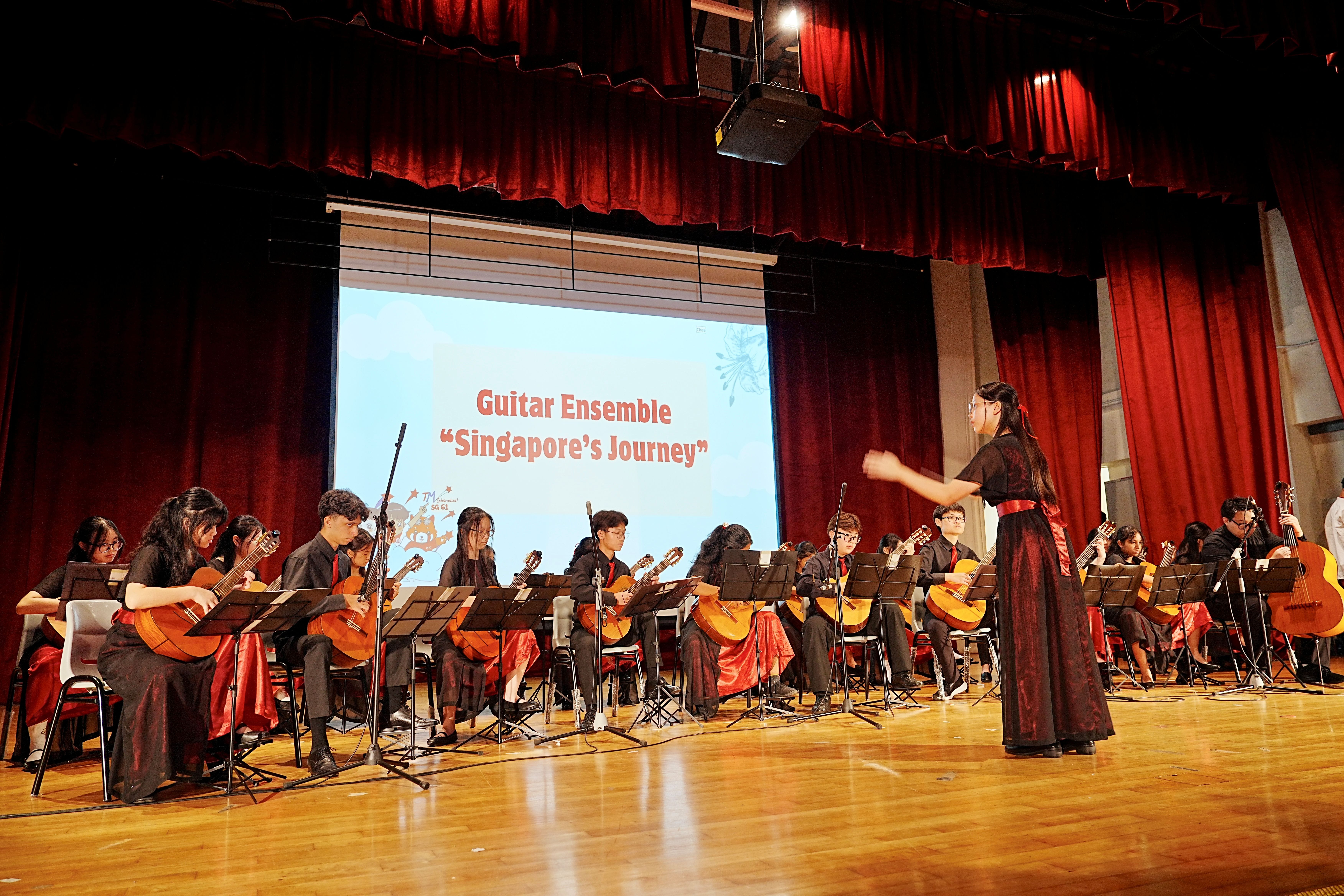 Students in black and red formal attire perform on stage under a projected slide reading "Guitar Ensemble – Singapore's Journey", led by a conductor.